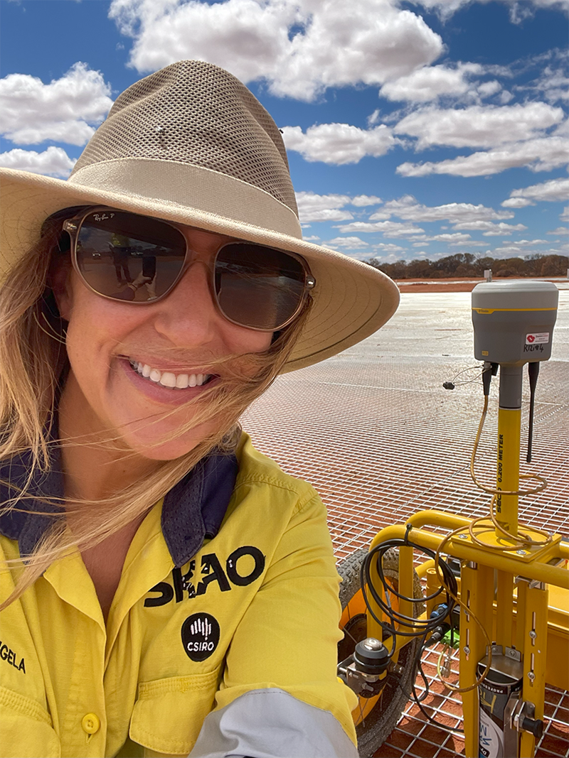 A brunette woman, Angela Teale, takes a smiling selfie wearing a yellow high-vis shirt with the SKAO and CSIRO logos on her left chest in front of a red soil and steel mesh on red Wajarri Country. Next to Angela is a small yellow robot with a tall pole holding a camera.  