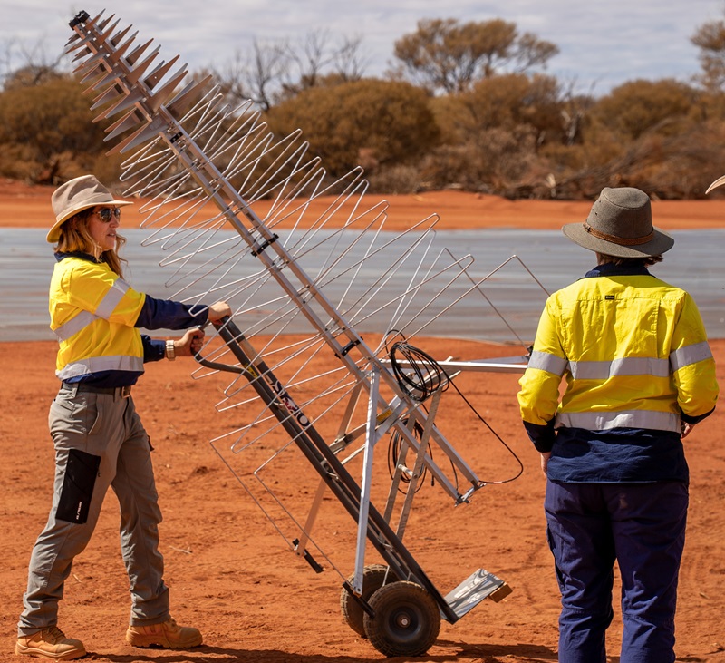 Angela stands wearing yellow and navy high vis shirt and sturdy boots on bright red Wajarri Country holding the handle of a trolley that balances a large silver metal tree-shaped antenna. 