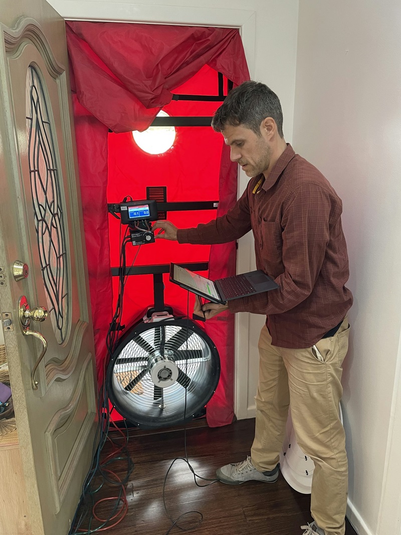 A man stands inside a house with a laptop by the front door. The door is covered in a red sheet with an exhaust fan at the bottom.