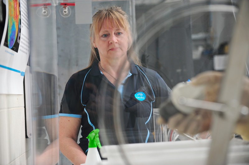 Dr Jane Hodgkinson in a blue CSIRO shirt, stands next to a dust chamber in a lab. In the forefront, there is a hand of someone working with items inside the clear dust chamber.