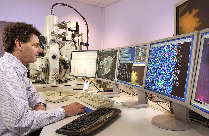 A man sits at a desk with 5 computer screens in front of him, each with different data and images. In the background is a large piece of equipment used for analysing samples.