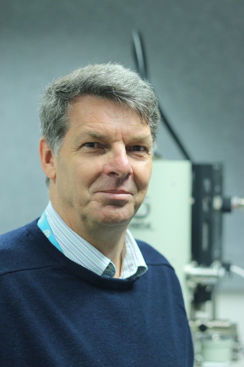 Colin MacRae in a blue jumper smiles at the camera. Behind him is a large piece of equipment