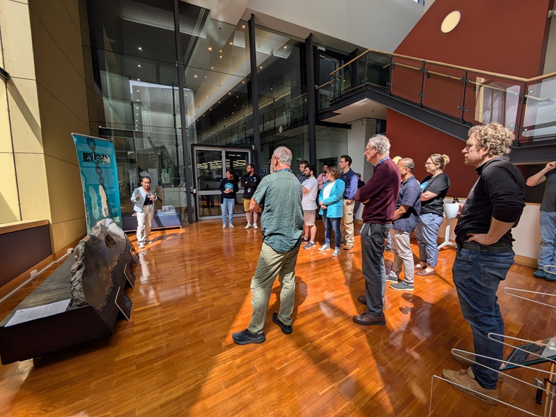 A group of people stands in a well-lit atrium at CSIRO Kensington, attentively listening to a speaker near the Bikpela spire, which is displayed on a wooden stand with informational posters in the background.