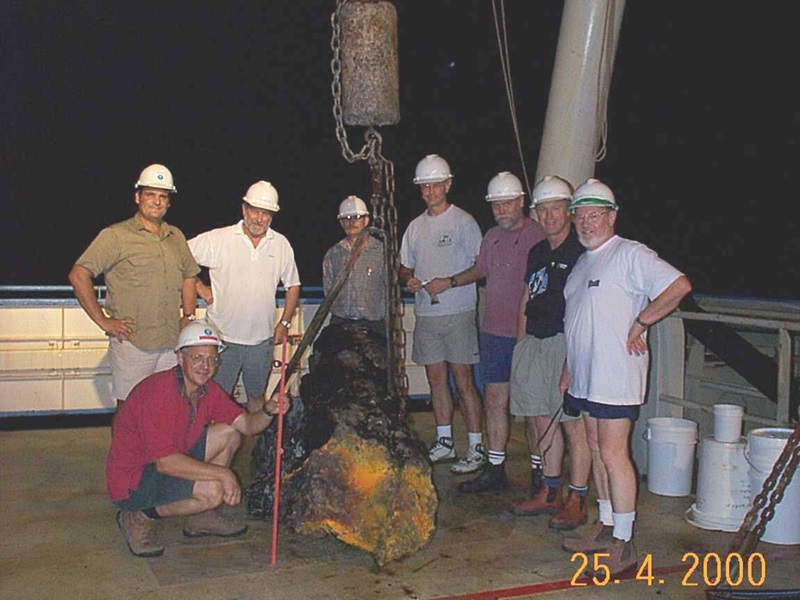 Dr Ray Binns, wearing a white hard hat and shirt, stands with his team on the deck of a ship, smiling as they secure the Bikpela spire with straps. The spire, a dark and rugged hydrothermal chimney with a bright yellow-orange sulphide deposit, is suspended mid-air by a crane. Other team members work nearby, wearing hard hats and casual attire.