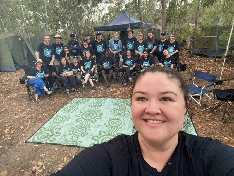 The 2024 CSIRO Garma Delegation smiling, wearing matching shirts, pose for a selfie at a campsite surrounded by tents and trees.