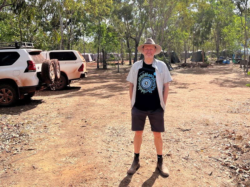 CSIRO Chief Executive Doug Hilton stands in the bush on red dirt surrounded by trees and white 4WDs.