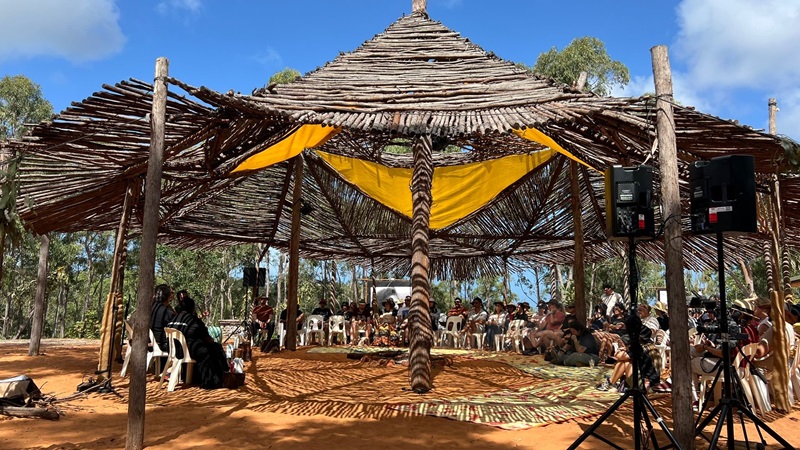 A gathering of people seated under a large, open-sided shelter made of wooden poles and branches.