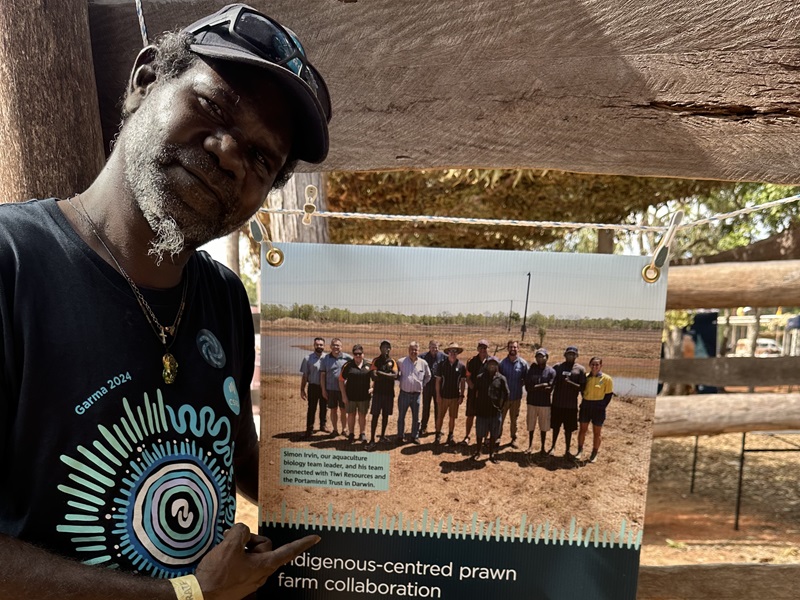 Ron stands next to a poster featuring a group photo of people, with text highlighting the Indigenous-centred prawn farm collaboration.