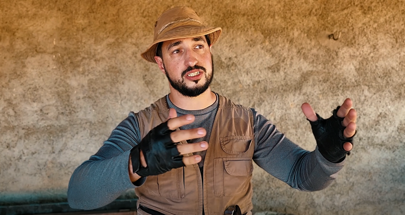 Vinnie is in front of a large rock wall, he wears clothes suitable for field work, including a vest with lots of pockets, fingerless gloves and a worn hat. Vinnie is holding his arms up an open as if presenting to a group of people