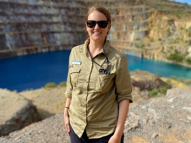 Person in khaki shirt wearing sunglasses standing with a quarry behind filled with turquoise blue water