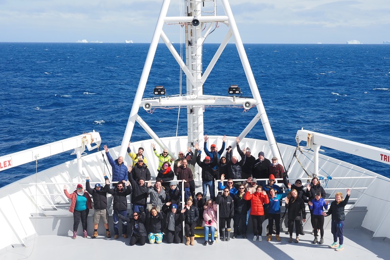 A group of people stand on the bow of a white ship at sea with icebergs visible on the horizon behind them.