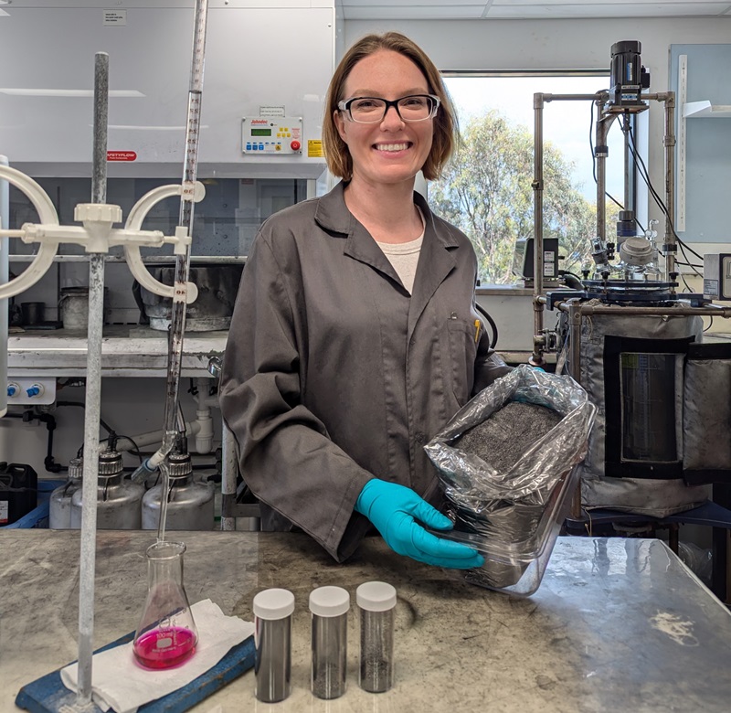 Kate wears a grey lab coat. She is standing in the lab and holding a container of fine black dust