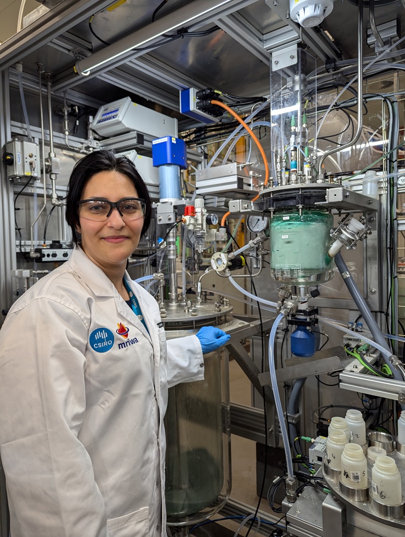 Neda wears a white lab coat and stands in front of a room sized piece of lab equipment