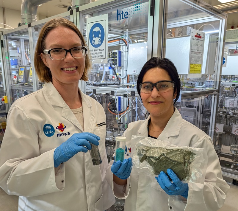 Kate and Neda wear white lab coats and blue gloves. They stand together smiling and hold small vials with lids of different coloured powders