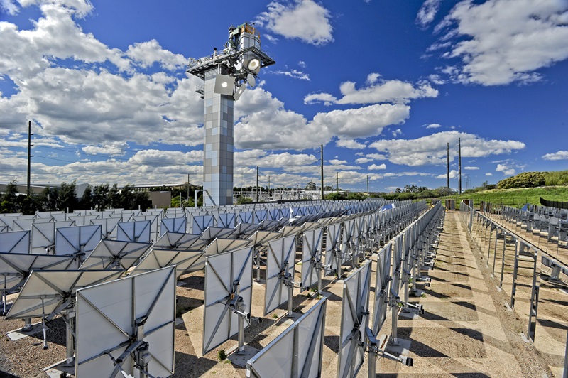An expansive array of solar panels in neat rows with a bright blue sky above. The panels are reflecting some of the fluffy clouds in the sky.