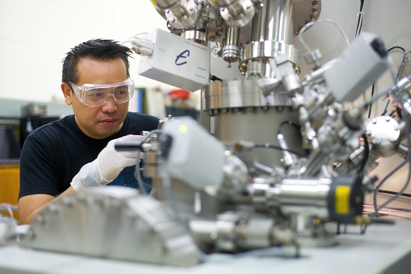 A scientists wearing safety goggles, gloves, and a navy blue t-shirt operates a complex looking machine to characterise the chemistry of a quantum device. 