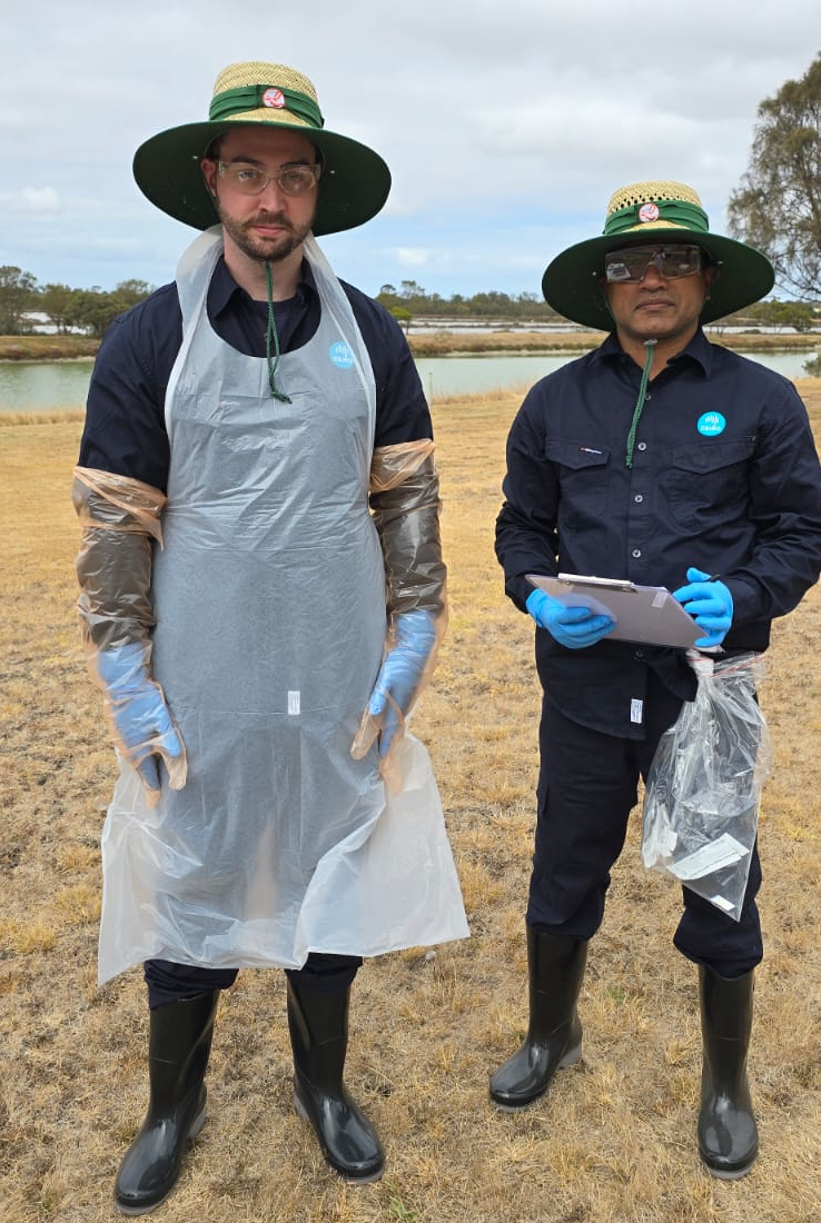 Two pwople standing next to each other in a field. One is wearing personal protective equipment including a plastic apron, gloves, glasses and rubber boots. The other is in work clothes, gloves, glasses and rubber boots. Both wear sun hats.