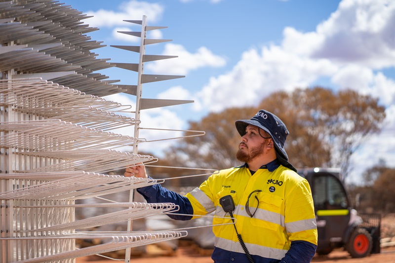 A man, Lockie, reaches for one part of a tall silver-metal tree-shaped SKA-Low antenna from a large stack under a cloudy blue sky with blurred construction vehicles in the background. Lockie wears high-vis protective clothes with the SKAO and CSIRO logos and his name embroidered on the chest.