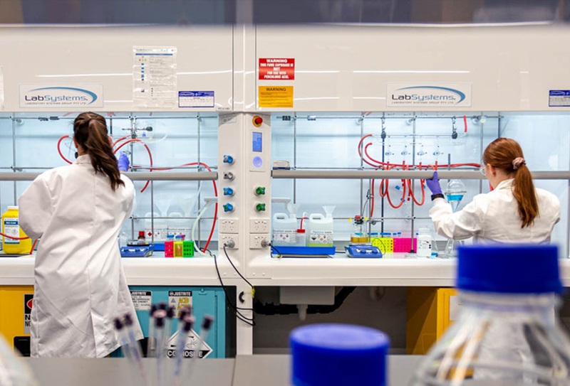 Two scientists are working in a lab, they are both seen from behind. The scientists are wearing white lab coats and have their long hair tied back.