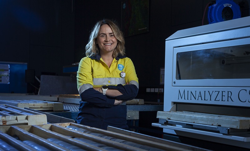 Renee wears an orange hi-vis shirt and stands in front of large piece of equipment used in the drill core lab. 