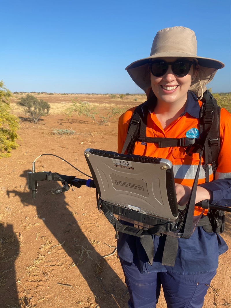 Jo is standing on red dusty earth. She wears a hi-vis orange shirt and hat, and holds a long scanning device (like a wand) in one hand. A tablet or imaging screen is harnessed to her chest like a forward facing backpack.