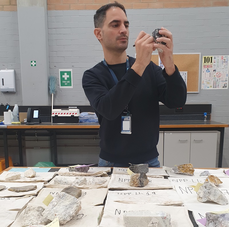 Mario stands at a table covered with rock samples, each with a label. Mario is holding one sample and writing on its label.