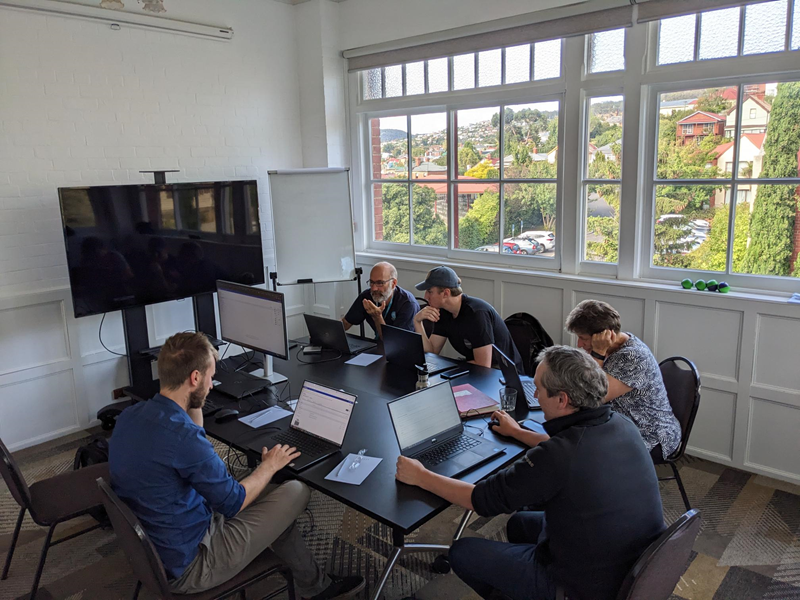 Five people sit at a table working on their laptops. In the background is a bank of windows with views to houses and hills.