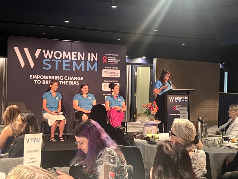 Three women in light blue shirts sit and a woman in a light blue shirt stands at the podium on stage.