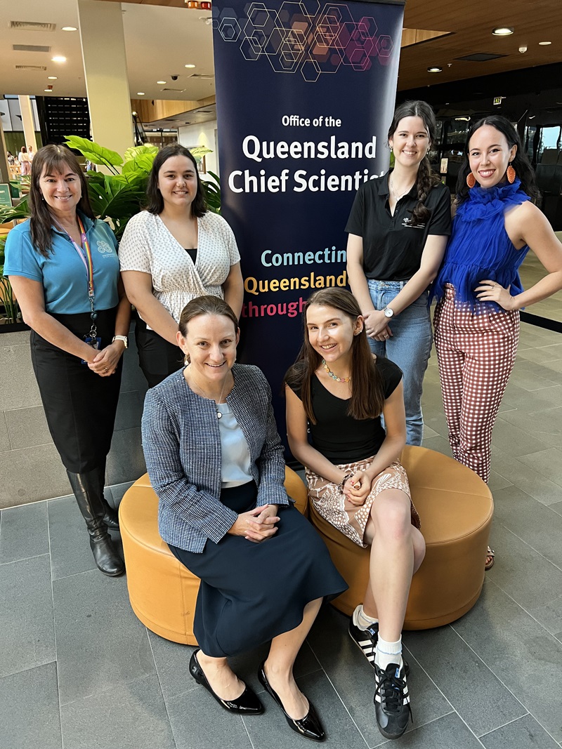 Four women stand and two sit in an office in front of a banner that reads 'Office of the Queensland Chief Scientist'.