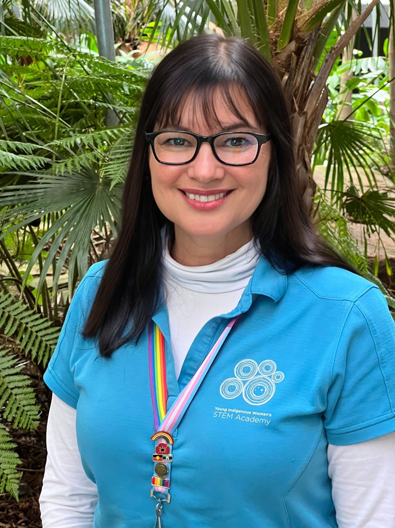 A woman in a light blue shirt and black reading glasses poses in front of a green tree.