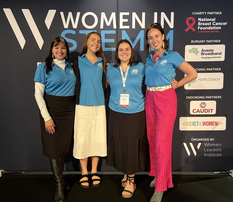 Four women wearing light blue shirts pose in front of a sign that says 'Women in STEMM'.