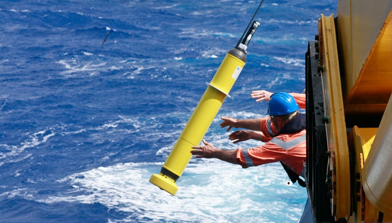 A yellow robotic instrument is lowered into the ocean by two men in orange hi-vis jackets.