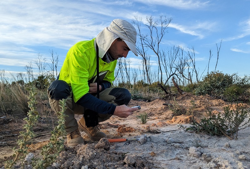 Person in high viz and a hat crouched on a scrubby rock with tools examining the rock