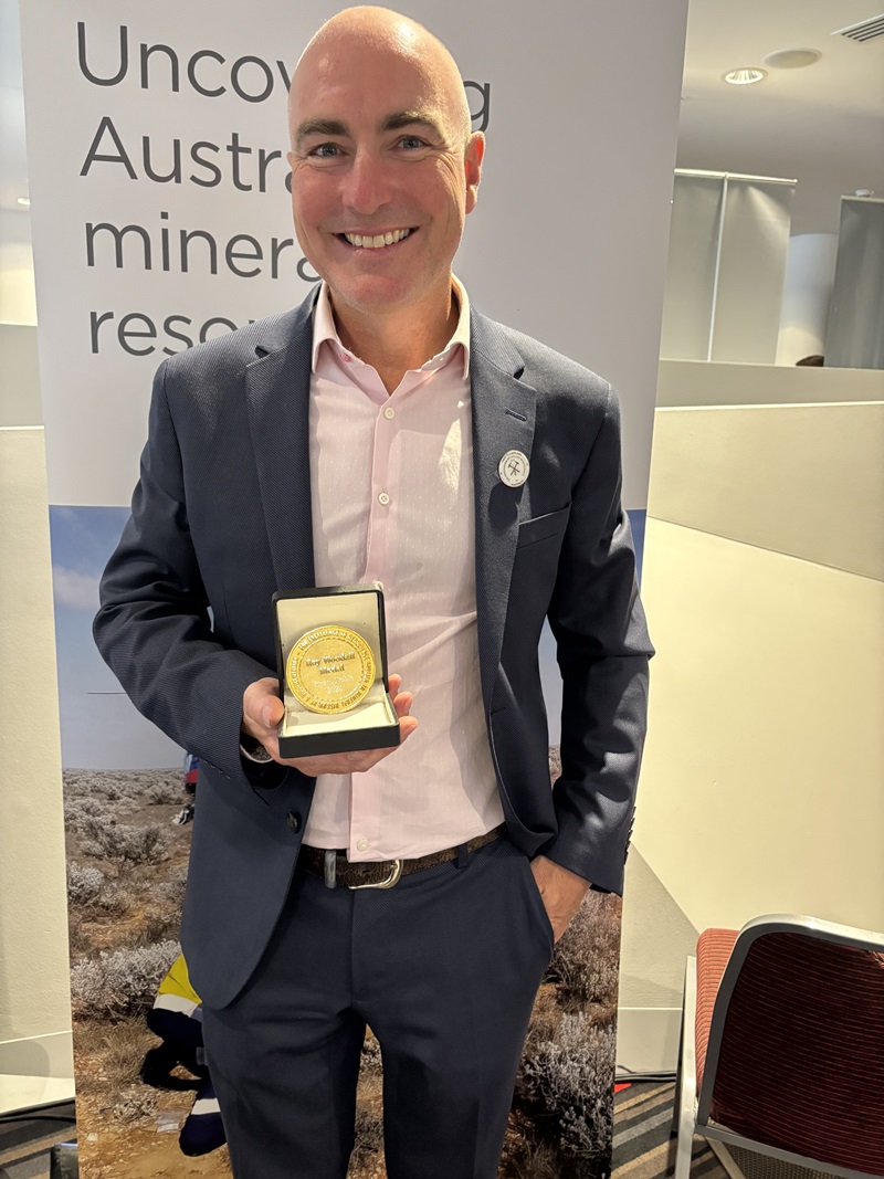 Ryan Noble in a blue suit smiles and holds out a gold-coloured medal in a black presentation box