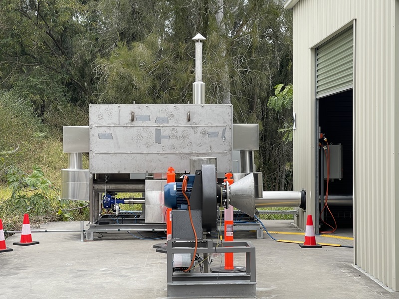 A truck-sized piece of equipment with an exhaust chimney, wires and control boxes. It sits in an industrial yard next to a large shed.