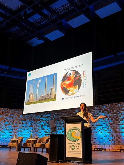 Ana Bugnot stands on a large stage, behind a podium with the AMSA signage, speaking to the audience. In the background is a large screen, suspended from the ceiling showing a slide of powerplant and globe.