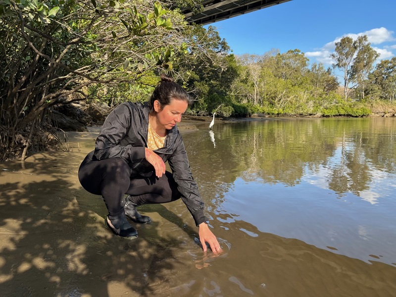 Ana Bugnot is crouched down on the edge of mudflats, reaching out touching the river water and sediment. A crane can be seen a few meters behind her, wading in the river. 