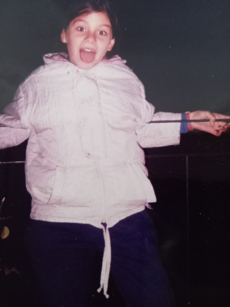 Eight-year-old Ana Bugnot wearing a pink puffer jacket leans against a railing smiling at the camera with her mouth open and her tongue poking out.