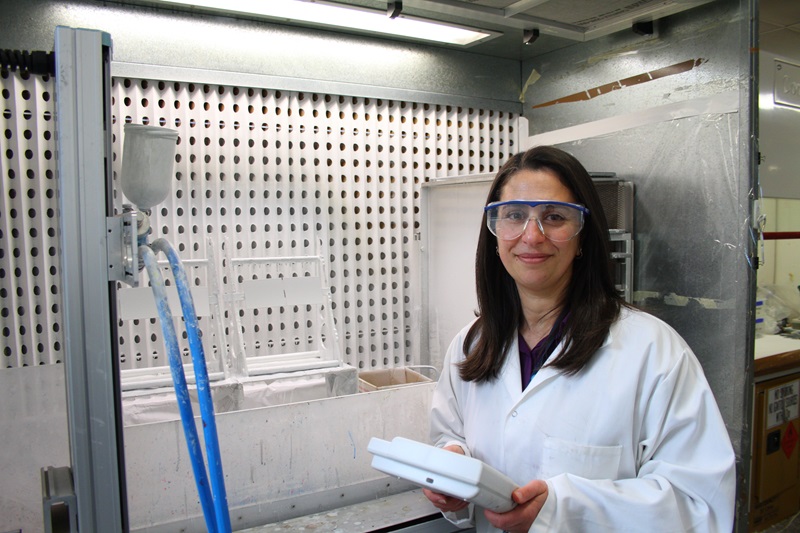 CSIRO researcher Ranya Simons stands in the CSIRO paint lab wearing a white coat and eye protection, smiling at the camera.