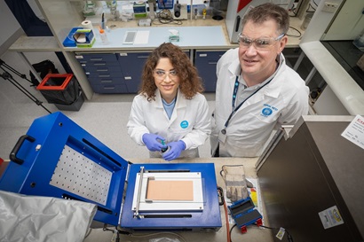 A male and female scientist both wearing lab coats and safety goggles looking up at the camera from a lab bench. 