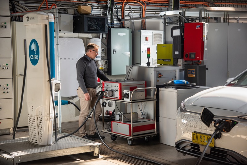 A CSIRO energy scientist, wearing a white lab coat and safety glasses, stands in front of a white electric vehicle (EV) in a brightly lit laboratory surrounded by technnical equipment