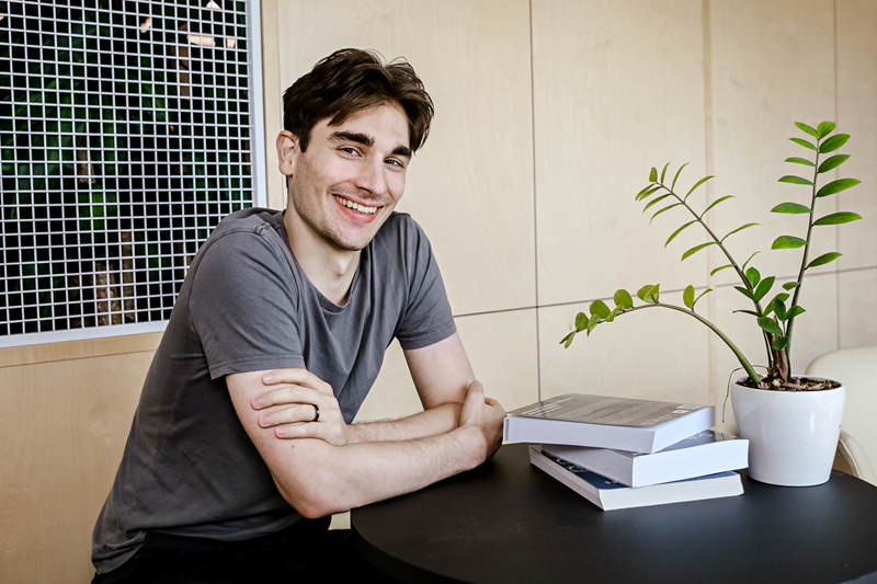 Aaron Nicolson sits at a desk, smiling at the camera. Aaron has short dark hair and wears a grey t-shirt. The desk is black, and on it is a stack of three books, and a green leafy plant in a white pot.