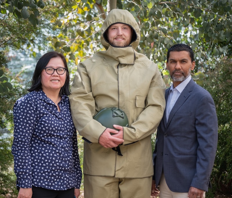 Three people standing together. The one in the centre is wearing a military style uniform with a hood. He is holding a military helmet in his hands. 