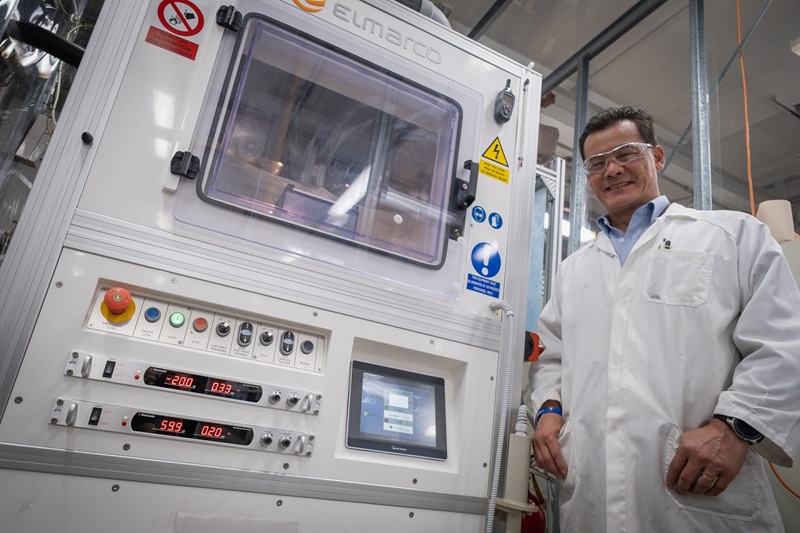 A man dressed in a white lab coat and safety glasses  standing beside a large technical-looking machine with a number of buttons, switches and lights on its face. 