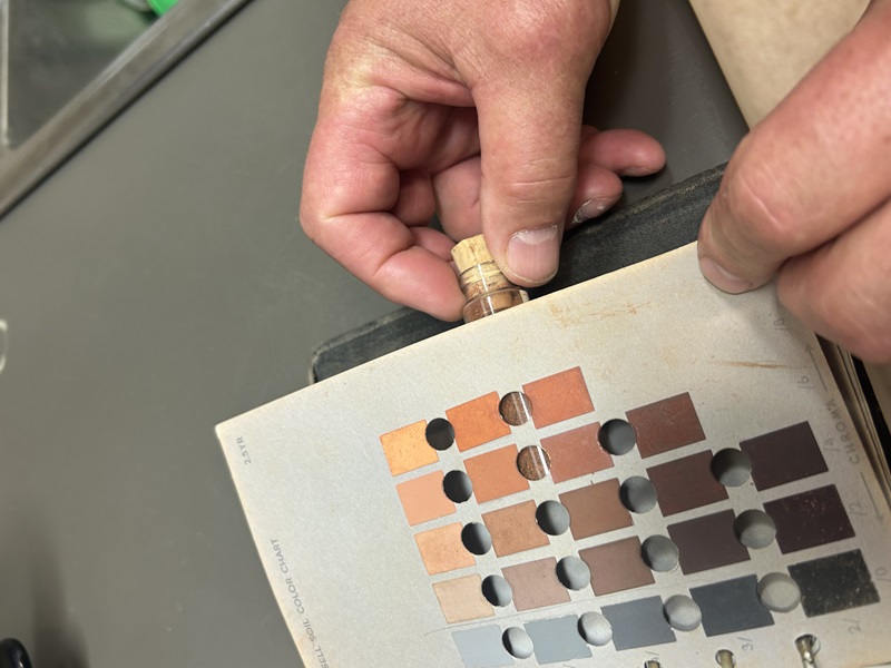 A close-up image showing two hands holding a soil sample and comparing its colour against a Munsell Soil Color Chart. The chart displays various shades of brown and reddish tones, with circular cutouts for easy soil colour matching.