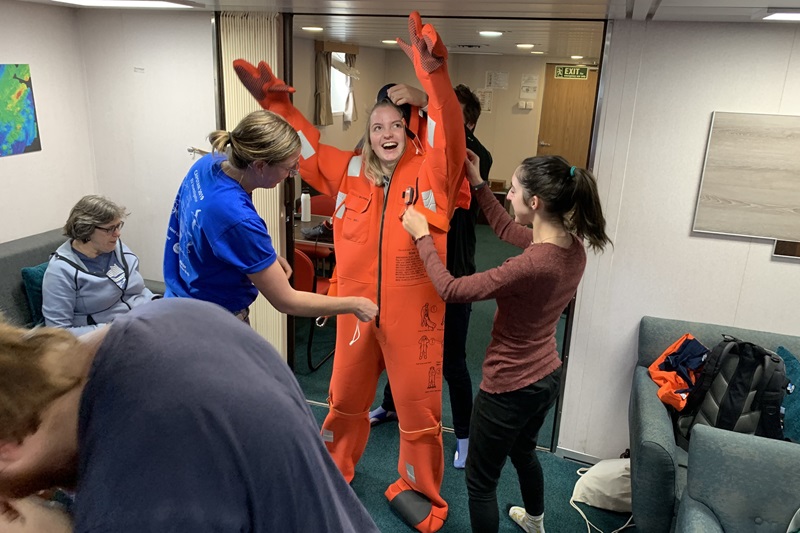 A group of people help another person put on a bright orange safety suit.