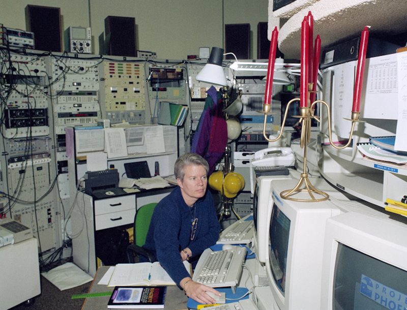 Woman with grey hair and wearing blue uniform sitting in a room crowded with computers.