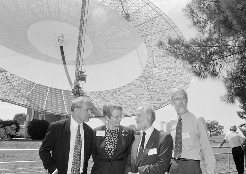 Three men and a woman standing under a tree in front of a large radio telescope.