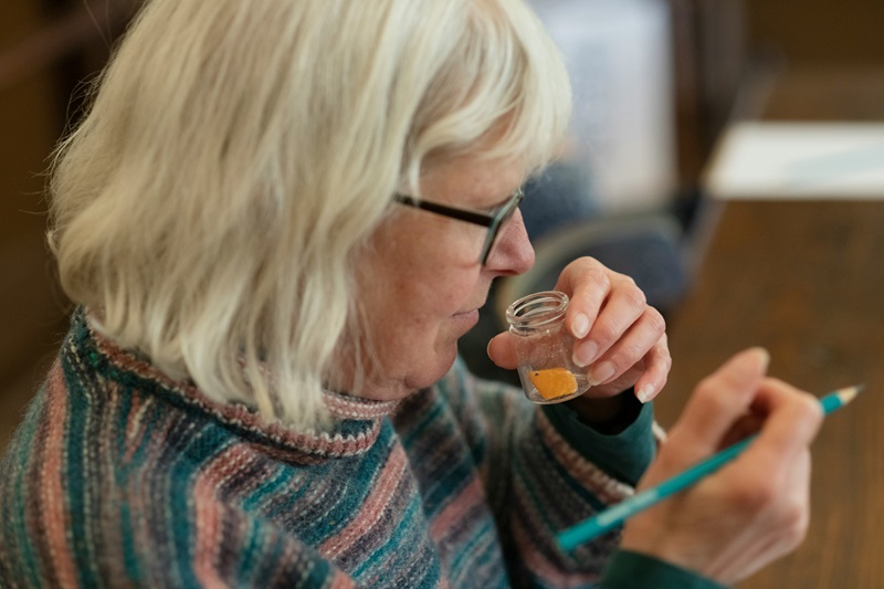 An older woman smells a piece of orange in a glass vial while holding a pencil.