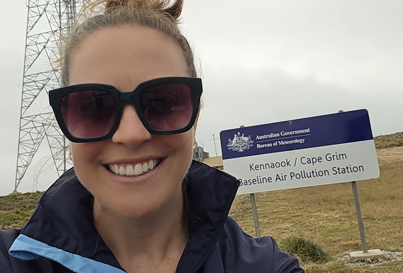 A woman wearing sunglasses smiles in front of a road sign that reads 'Kennaook Cape Grim Baseline Air Pollution Station'.